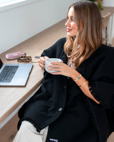 Woman sitting at a desk holding a mug with a laptop open in front of her wearing a warmur office chair blanket